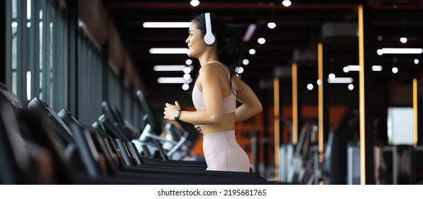 Panoramic Side view of beautiful young asian woman running on treadmill and listening to music via headphone with arm a smart watch for tracking speed during sports training in a gym. - Powered by Shutterstock