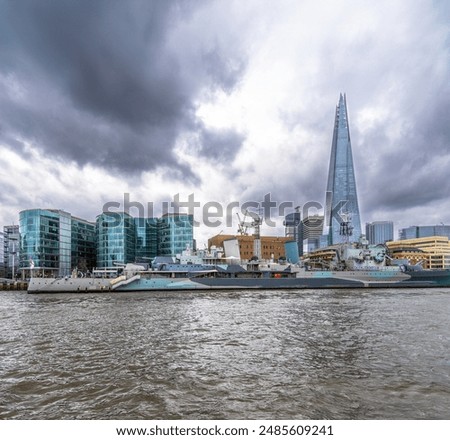 Panoramic side view of the floating war museum ship HMS Belfast docked on the River Thames with The Shard skyscraper in the background under a rainy cloudy sky.