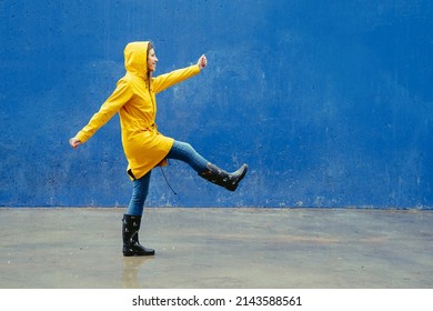 Panoramic Side View Of Woman Wearing A Yellow Raincoat Walking Funny On Blue Wall. Full Length Body Of Woman Under The Rain With Wellingtons And Blue Background. Seasonal Concept Winter And Spring.