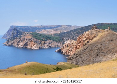 Panoramic summer view of the dramatic coastal landscape of the bay, featuring rugged cliffs, the sea, and the ruins of a fortress on the hilltop.