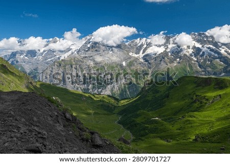 Panoramic summer view of green alpine valley and rugged snowcapped mountains under blue sky near Mürren, Switzerland.