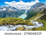 Panoramic summer view over a cascading mountain stream and rocky alpine meadow down to the turquoise Schlegeis Stausee reservoir, surrounded by steep snowcapped peaks and forests in the Zillertal Alps near Mayrhofen, Tyrol, Austria. Bright blue sky with clouds above the dramatic high mountain valley landscape.