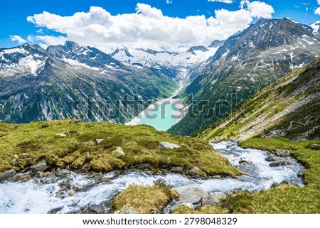 Panoramic summer view over a cascading mountain stream and rocky alpine meadow down to the turquoise Schlegeis Stausee reservoir, surrounded by steep snowcapped peaks and forests in the Zillertal Alps near Mayrhofen, Tyrol, Austria. Bright blue sky with clouds above the dramatic high mountain valley landscape.