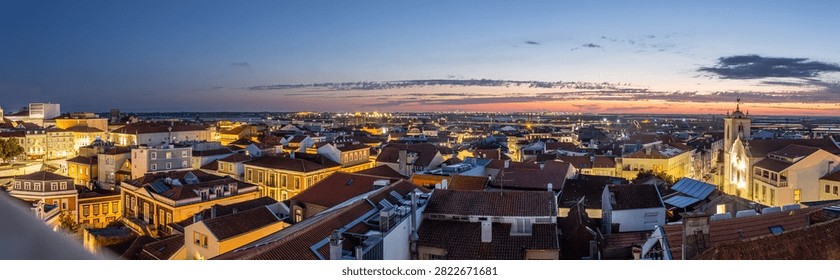 Panoramic sunset view over the rooftops of Aveiro, Portugal, captured from a high vantage point. The illuminated historic center, church steeples, salt marshes, and Aveiro lagoon create a stunning cityscape during the blue hour.