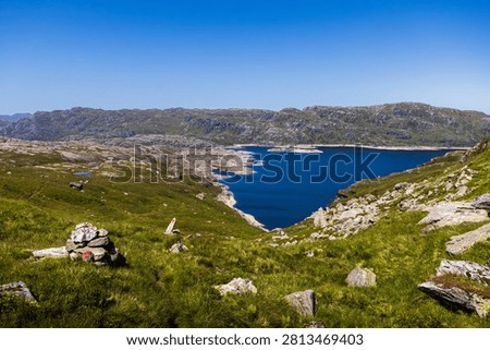A panoramic view across the deep blue waters of Lykilsvatnet from the mountain plateau of Etnefjellet in Etne, Vestland, Norway. Rocky highlands, hiking trail markers, and expansive alpine scenery showcase the wild beauty of Western Norway