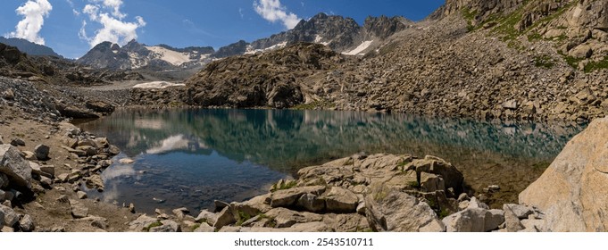 Panoramic view of alpine lake surrounded by rocky terrain and reflecting snow-capped peaks under blue sky. Water mirrors the rugged mountains, capturing tranquil beauty of high-altitude landscape - Powered by Shutterstock