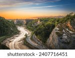 Panoramic view of Avon Gorge near center of city of Bristol, England, in summer at sunset; cliffs on both sides, highway with moving cars on bottom and sunset sky in background