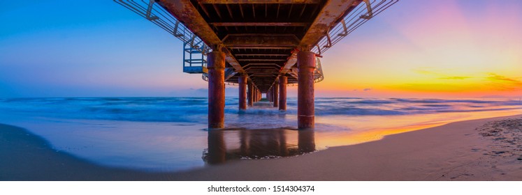 Panoramic view of beach sunset under a pier with lines leading to vanishing point