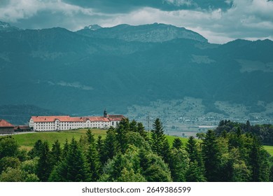 Panoramic view of a Benedictine monastery surrounded by pine trees with the Swiss Alps in the background, cloudy summer day, nature and culture in harmony.