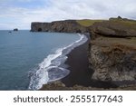 Panoramic view of a black volcanic beach with rocky cliffs, waves crashing on the shore, and a partly cloudy sky, creating a dramatic atmosphere.