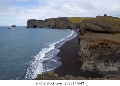 Panoramic view of a black volcanic beach with rocky cliffs, waves crashing on the shore, and a partly cloudy sky, creating a dramatic atmosphere. - Powered by Shutterstock