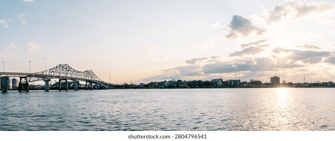 A panoramic view of a bridge spanning over a river at sunset, with a city skyline in the distance under a partly cloudy sky.