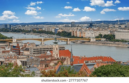 Panoramic view of Budapest features the Danube River, the yellow Margaret Bridge, and a cityscape of historic architecture under a bright blue sky with fluffy white clouds.
