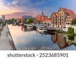 Panoramic view of the Bydgoszcz waterfront at sunrise with boats along the Brda River, historic half-timbered buildings, and modern structures in Poland