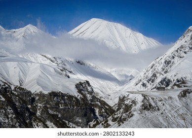 A panoramic view captures the rugged, snow-covered peaks and deep valleys of the Greater Caucasus Mountains in winter, Georgia. A winding mountain road is visible in the foreground, leading towards a small building perched on a snowy ridge under a vibrant blue sky.