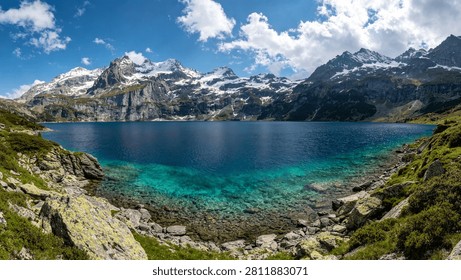 Panoramic view of a clear turquoise alpine lake surrounded by rugged rocky slopes and snow-covered mountain peaks beneath a bright blue sky with white clouds. Scenic wilderness landscape suitable for travel, adventure, nature, and environmental themes.