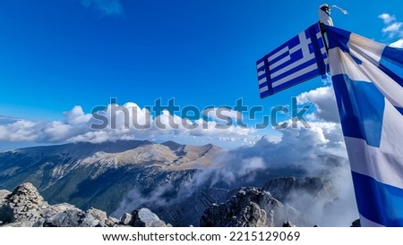 Panoramic view from cloud covered mountain summit of Mytikas Mount Olympus, Mt Olympus National Park, Macedonia, Greece, Europe. Greek national flag waving in the wind on top. Mountain ranges