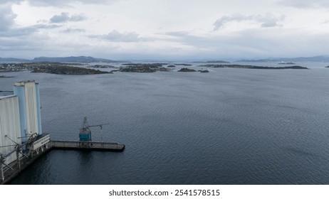 A panoramic view of a coastal landscape featuring calm waters, scattered islands, and a harbor with a crane. The sky is overcast, adding a dramatic effect to the scene. - Powered by Shutterstock