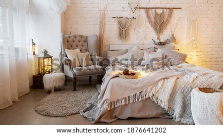Panoramic view of cozy bedroom with crafted home decor over bed, armchair and stool in apartment with white interior design in boho chic style