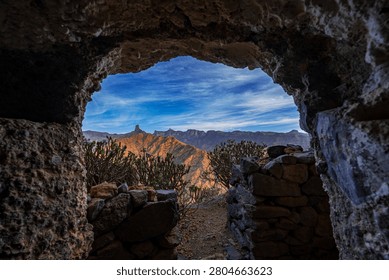 Panoramic view of the dramatic mountain landscape of Gran Canaria framed by the entrance of a historic cave dwelling. The rugged volcanic peaks, native vegetation, and iconic rock formations create a unique perspective on the natural beauty and cultural heritage of the Canary Islands, Spain.