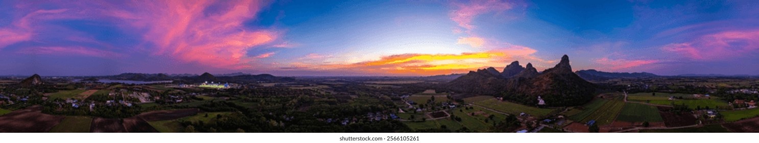 A panoramic view of dramatic mountain peaks silhouetted against a vibrant sunset sky, with fiery hues of red, orange, and pink blending into the fading blue. sunflowers fields around the mountains.