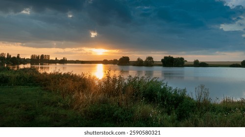 Panoramic view of Dramatic Sunset Over a Serene Lake. Dark clouds hang over the lake, creating a contrast with the warm glow of the setting sun reflected in the water.