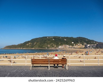 Panoramic view of an elderly couple sitting with their backs turned on a bench in the middle of a promenade in front of the crowded beach in the tourist city of San Sebastian in the Basque Country - Powered by Shutterstock