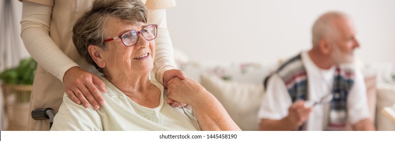 Vista panorámica de una anciana en silla de ruedas sosteniéndose de la mano con el voluntario que la apoya parado detrás de ella
