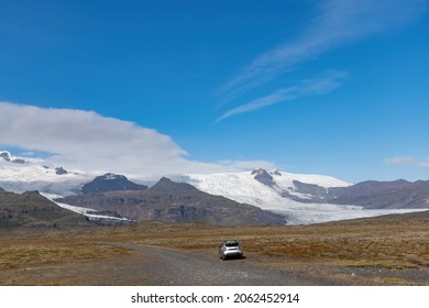 Panoramic view of the Fjallsarlon glacier in Iceland seen from a dirt road close to the terminus of the glacier with a car parked on the road against a blue sky with some feather clouds