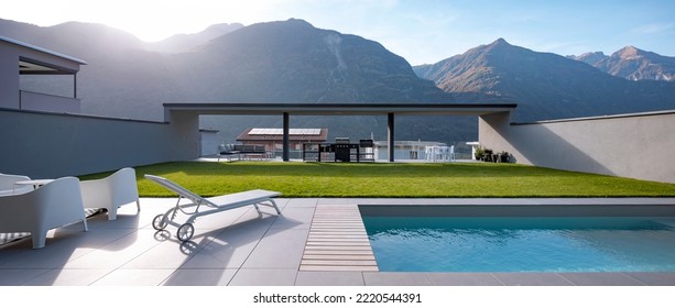 Panoramic view garden with pool of modern villa. There is a sitting area with armchairs, a deck chair, and at the back is the barbecue. In the background there are mountains. Banner, nobody inside