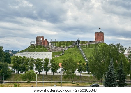 Panoramic view of Gediminas Tower and the Upper Castle ruins situated on Castle Hill in Vilnius, Lithuania. The iconic medieval red-brick tower stands prominently on the right side of the lush green hill, flying the Lithuanian national flag. To the left, the ruined remains of the castle walls and foundations are visible. A modern inclined elevator track runs up the grassy slope, providing access to the historic site. At the base of the hill sits the white Palace of the Grand Dukes of Lithuania with its distinctive crenellated roofline. The foreground features verdant trees, a road with a passing car, and pedestrian areas. The sky above is overcast with dramatic gray clouds. The scene captures a blend of Gothic architecture, natural landscape, and urban elements, representing one of Lithuania's most significant historical landmarks and a major tourist attraction.