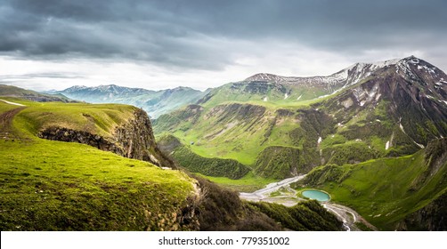 Panoramic View From The Grassy Hill On The Kazbek Mountains And The Green Valley Crossed With Roads And With A Blue Lake In Georgia Against Cloudy Sky
