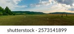 The panoramic view of the green field by the hills under the blue sky with clouds