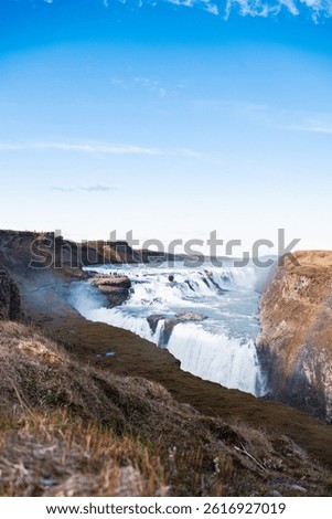 Panoramic view of Gullfoss waterfall cutting through rugged Icelandic cliffs, with turquoise waters and clear skies over the highland plateau.