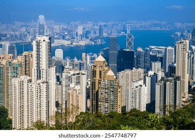 A panoramic view of Hong Kong dense cityscape, showcasing towering skyscrapers and a lush green hillside. The blue sky with a single fluffy cloud provides a picturesque backdrop. - Powered by Shutterstock