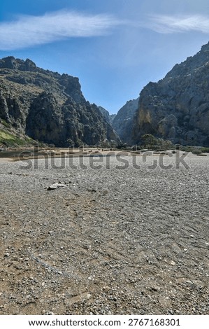 Panoramic view of the interior limestone canyon at Torrent de Pareis in Sa Calobra. Towering vertical cliffs surround a vast pebble riverbed under a bright blue sky. Tourism and nature concept.