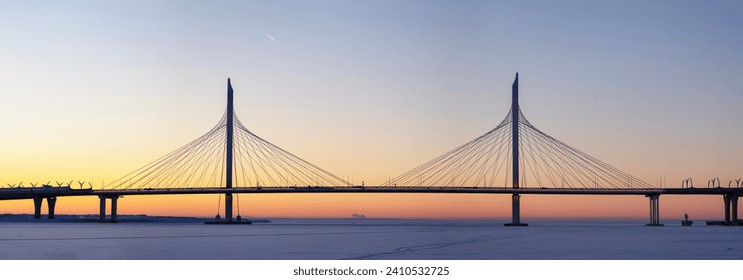 Panoramic view: Large cable-stayed expressway bridge at sunset over an ice-covered bay with an airplane flying high