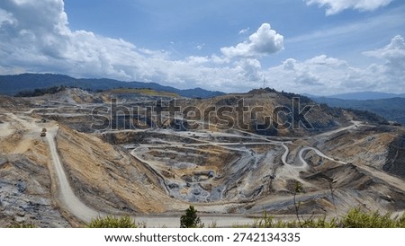 Panoramic view of a large open-pit tin mining operation showing terraced earth and haul roads in rural area of Perak, Malaysia. Expansive landscape of an active open-cast tin mine with heavy machinery and winding dirt roads against a mountainous backdrop. Wide perspective of extensive earthworks and excavation at a commercial mining site surrounded by natural forest.