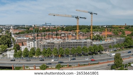 Panoramic view of large urban construction site with four cranes, scaffolding-covered buildings, residential homes in the background and road with traffic in front. Construction site in Kiel, Germany.