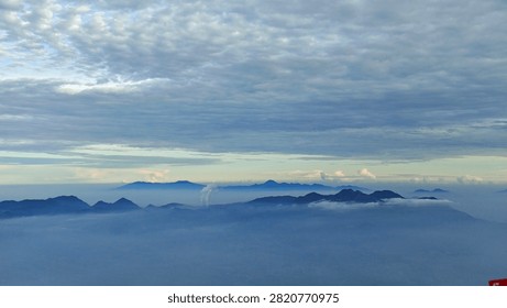 Panoramic view of layered mountain ranges beneath a dramatic cloudy sky with morning mist and soft atmospheric light. A peaceful natural landscape ideal for travel, adventure, environmental, and outdoor concepts.