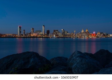 Panoramic view of the Liverpool skyline across the Mersey river at night, with pink and turquoise reflections in the water, as seen from the Seacombe, just across the river