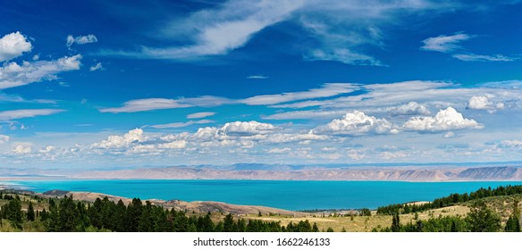 Panoramic View Looking Down Mountain With Green Trees And Grass At Bright Blue Lake With Mountains Beyond Under Blue Skies With White Clouds.