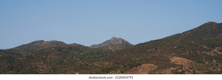 Panoramic view of a lush mountain range under a clear blue sky, with green valleys and a distant hilltop structure, showcasing the natural beauty of the landscape