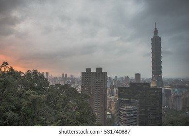 Panoramic view of majestic skyscraper and city skyline under a dramatic overcast sky at sunset, captured from a lush green hillside in Taipei, Taiwan.