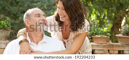 Panoramic view of a mature tourists couple sitting in a green garden during a sunny day on a summer holiday enjoying each others company, hugging and loving outdoors. Senior people lifestyle.