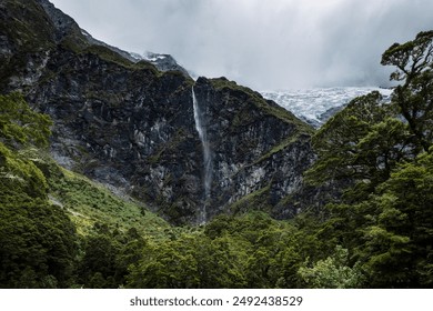 A panoramic view of a mountain and glacier with a cascading waterfall, surrounded by dense forest and lush greenery.