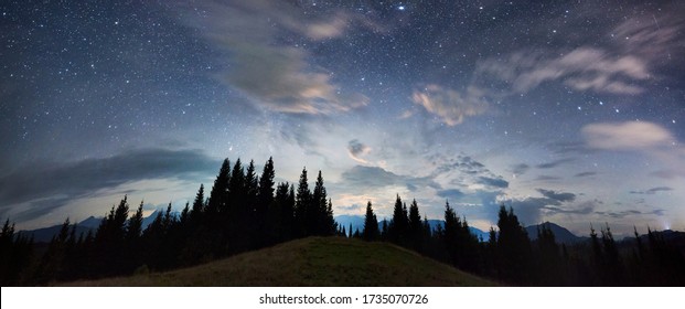 Panoramic View Of A Mountain Hill Facing Spruce Forest And A Mountain Ridge, Magical Starry Night Sky With Purple Clouds Above Beautiful Horizon, Copy Space. Concept Of Travelling, Galaxy And Nature.