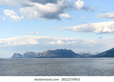 Panoramic view of mountain range across calm sea under cloudy sky. Wide panoramic vista of rugged mountain peaks rising above the open ocean. Fluffy white clouds fill the bright blue sky above the calm sea surface, creating a peaceful nordics-inspired coastal landscape.