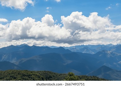 A panoramic view of a mountain range under a partly cloudy sky.  The mountains are dark blue-green, layered, and extend to the horizon. Lush green vegetation...