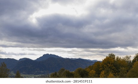 Panoramic view of mountain range under heavy overcast sky. Autumn forest in foreground, moody lighting with sun rays breaking through dramatic storm clouds, vast highland scenery.